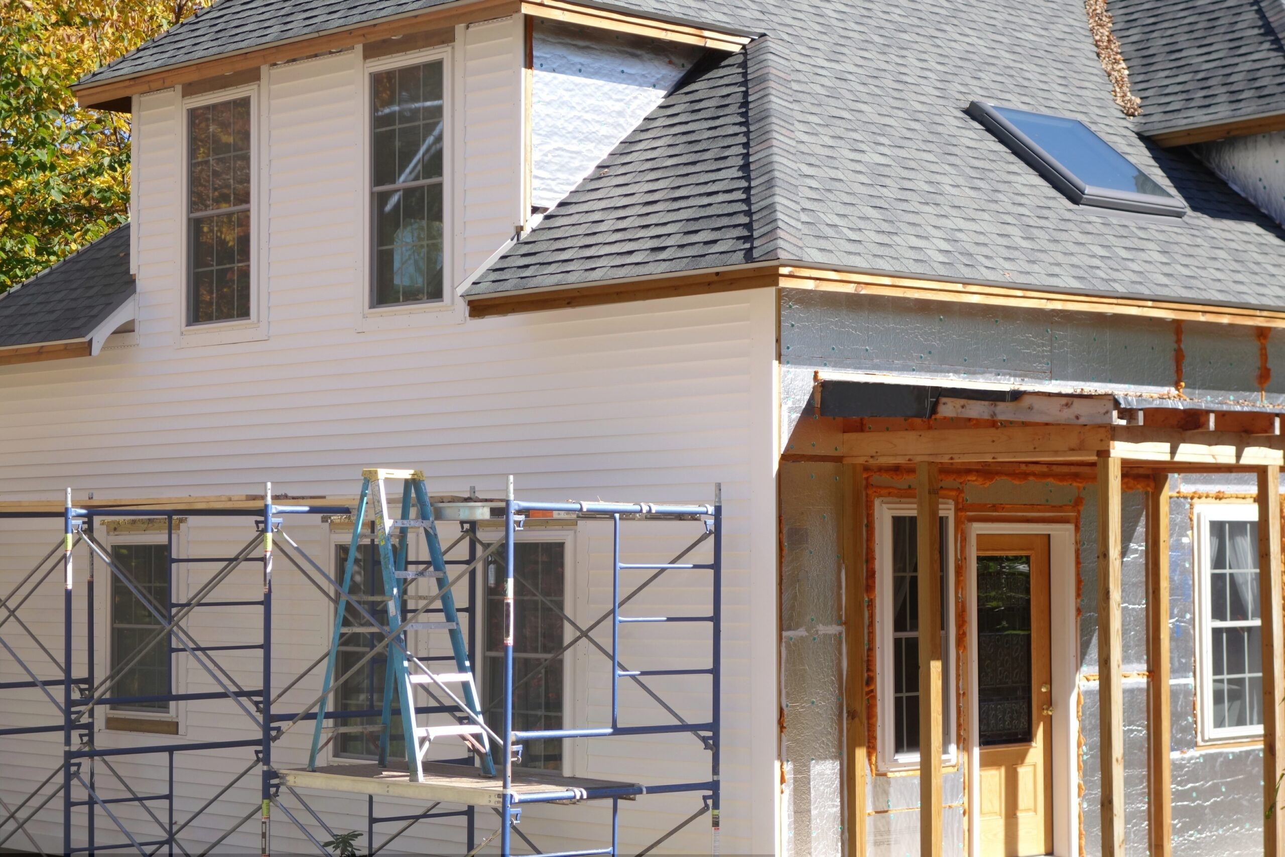 House exterior under renovation, featuring scaffolding, new siding, and unfinished areas. A doorway and windows are visible, highlighting ongoing improvements.