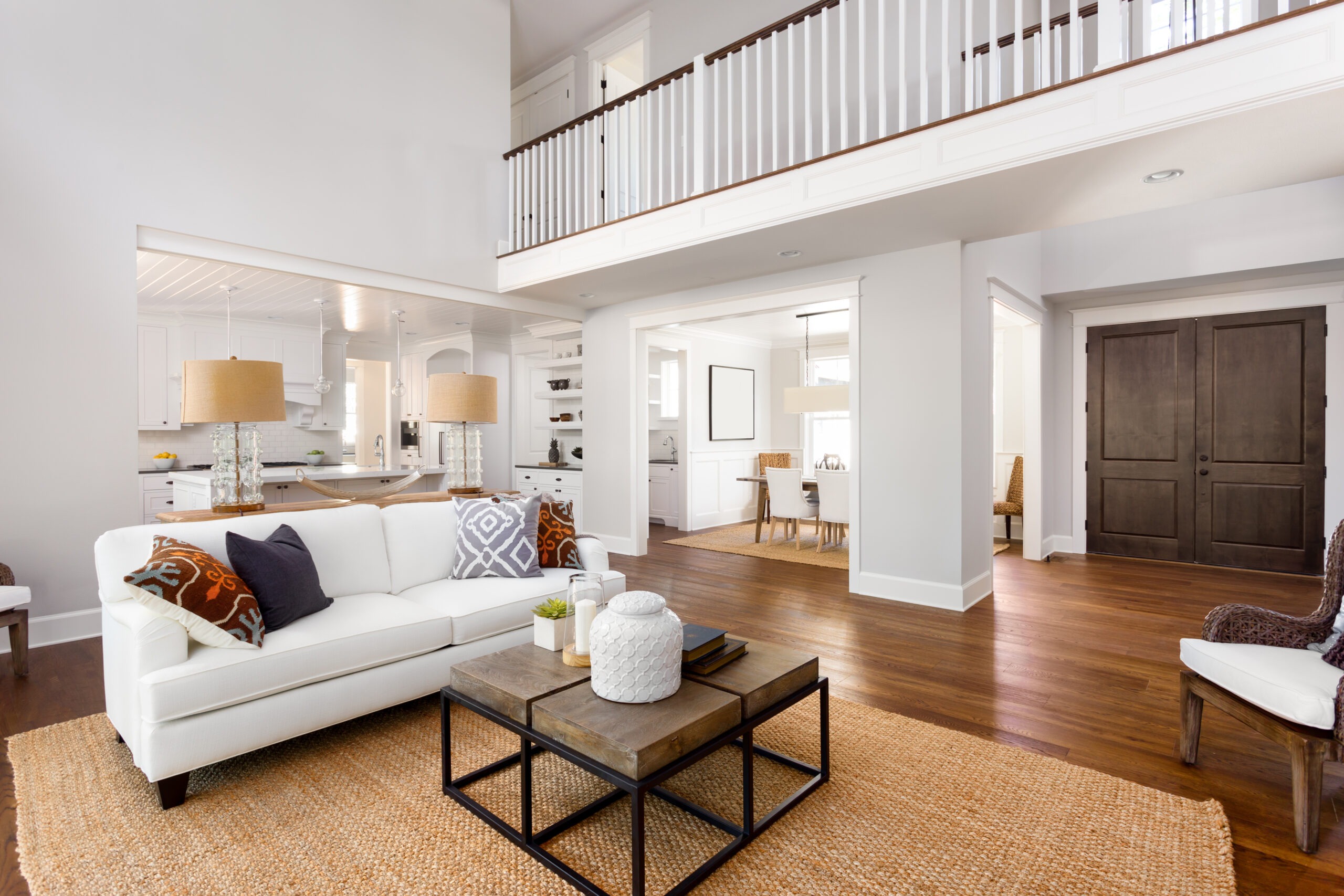 Bright and airy living room featuring a white sofa, patterned pillows, and a wooden coffee table. The open layout connects to a modern kitchen and dining area.